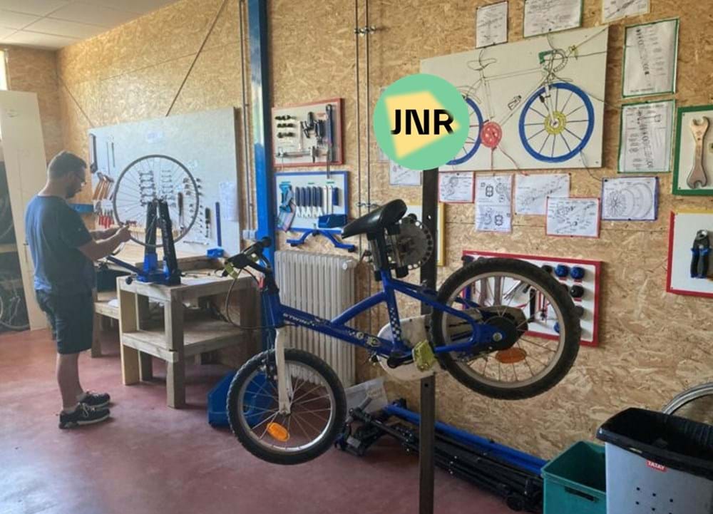 Un vélo enfant est suspendu à un pied d'atelier, tandis qu'un bénévole répare une roue à l'arrière-plan, dans la Vélobricolade de Roue Libre à Barberaz, au Percolateur. Le logo des Journées Nationales de la Réparation est également apposé sur l'image.